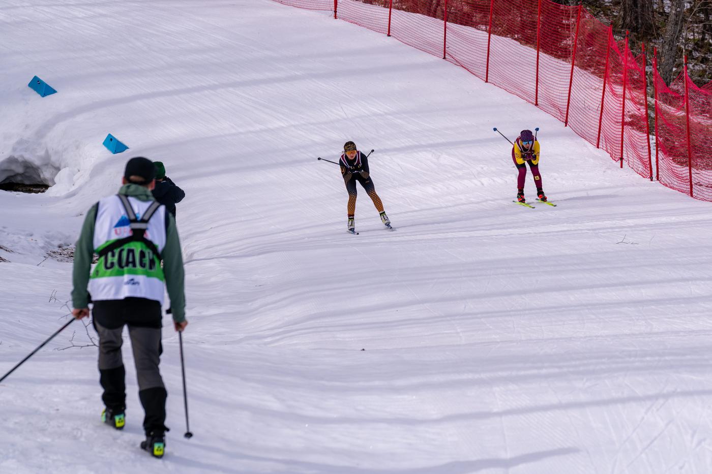 Greta Bochenek, CMU and Amaeli Kam-Magruder, St. Olaf, climb a hill during the 15 km freestyle race at USCSA National Championships in Lake Placid, with a coach watching from the course side.